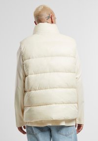 Cream-colored puffer vest with horizontal quilted sections, high collar, and a smooth shiny texture. Worn over a light shirt and jeans.