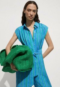 Donna che indossa un vestito senza maniche a righe blu-verdi, tiene in mano una grande coperta verde testurizzata davanti a uno sfondo bianco.
