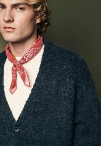 Dunkelblauer Strickcardigan mit strukturiertem Finish, ausgestattet mit einem V-Ausschnitt und einem schwarzen Knopf. Ein rotes Bandana mit weißen Mustern liegt um den Hals.