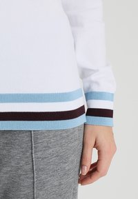 Witte top met lange mouwen en blauwe, bruine en lichtblauwe horizontale strepen op de manchetten, gedragen met grijze broek, waarbij een deel van een hand zichtbaar is.