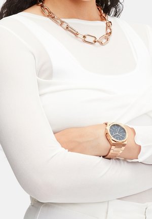 Femme portant un haut blanc à manches longues, un collier chaîne épais en or rose et une montre en or rose avec un cadran bleu foncé.