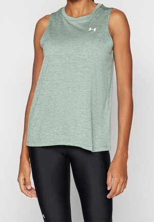 Person iført en ærmeløs lysegrøn træningstop og sorte tætsiddende leggings, stående med armene afslappet ned langs siden.