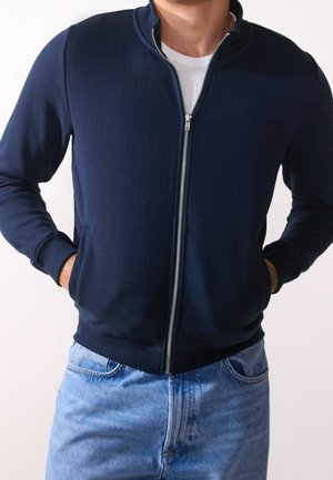 Navy zip-up sweatshirt made of cotton blend, featuring a high collar, front pockets, and ribbed cuffs. Paired with light blue jeans.