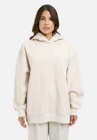 Eine Frau trägt einen lockeren, hellbeigen Hoodie mit fallenden Schultern und gerippten Bündchen und steht vor einem schlichten weißen Hintergrund.