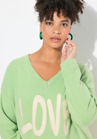 Pullover verde oversize con scollo a V e testo "LOVE" color crema. Il tessuto sembra morbido. La modella indossa orecchini a cerchio verdi.
