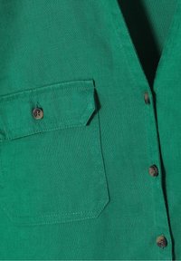 Section de chemise en tissu vert avec une poche à rabat boutonnée sur le côté gauche et des boutons foncés le long de l’ouverture devant.