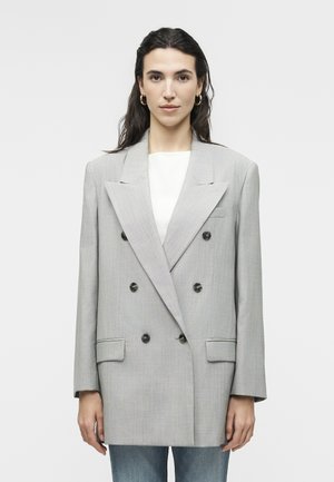 Vrouw met donker haar draagt een oversized lichtgrijze double-breasted blazer met zwarte knopen, een wit topje, gouden oorringen en een blauwe spijkerbroek.