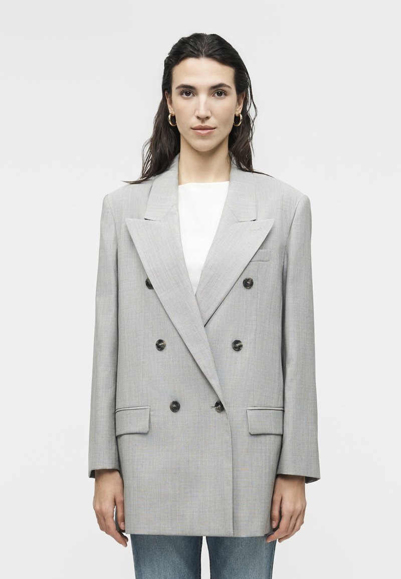 Γυναίκα με σκούρα μαλλιά φοράει υπερμεγέθη ανοιχτό γκρι σακάκι διπλής μπροστινής σειράς με μαύρα κουμπιά, λευκό τοπ, χρυσούς κρίκους σκουλαρίκια και μπλε τζιν.