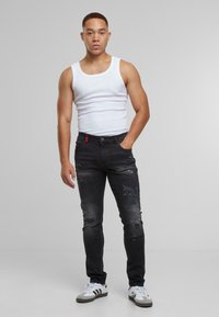 Noormees seisab üks käsi taskus, kandes valget tank topi, kulunud musti teksaseid ja valgeid jooksujalatseid mustade triipudega.