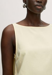 Hellgelbes ärmelloses Oberteil mit weiter Ausschnittform, glatter Textur und minimalen Nähten, ergänzt durch silberne Creolen.