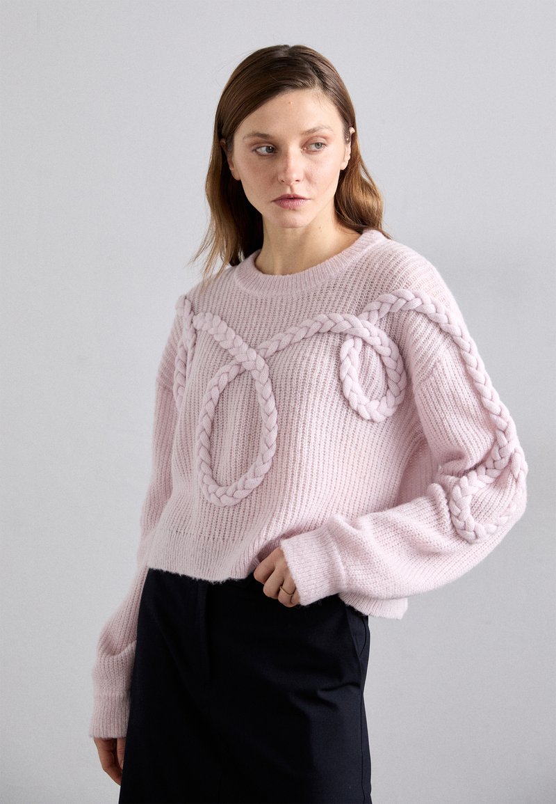 Pulover tricotat de culoare roz deschis, cu detalii împletite pe piept și mâneci. Textură cu nervuri și design scurt. Pantaloni negri vizibili dedesubt.
