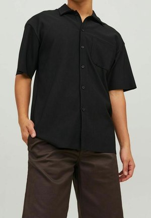 Mann trägt ein schwarzes Kurzarmhemd mit Brusttasche, dazu dunkelbraune Hosen, steht mit einer Hand in der Hosentasche.