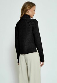 Schwarzer gerippter Rollkragenpullover mit lässigem Schnitt und hohem Kragen. Der Saum ist leicht gekürzt und zeigt plissierte, hellfarbene Hosen.
