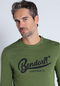 Long-sleeve olive green t-shirt made of cotton, featuring a prominent black logo "Bendorff CONTRASTS" across the chest.