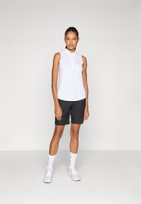 Hvit ermeløs polo skjorte med krage, kombinert med svarte shorts. Modellen har på seg hvite sokker og lyse grå treningssko med røde detaljer.
