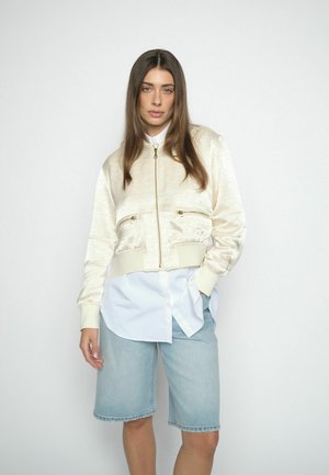 Kvinde med langt brunt hår iført cremefarvet satin bomberjakke, hvid skjorte med knapper og lyseblå knælange denimshorts foran en ensartet baggrund.