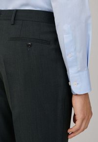 Donkergroene broek van textured stof, met een achterzak met een zwarte knoop. Lichte blauwe manchetten van een shirt zichtbaar met knopen.