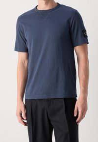 Mies, który ma na sobie prostą ciemnoniebieską koszulkę z krótkim rękawem z czarną naszywką logo na lewym rękawie oraz czarne spodnie.