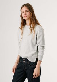 Maglione grigio chiaro con colletto a giro, polsini a coste e un sottile dettaglio testuale sull'orlo, abbinato a jeans blu scuro con doppi bottoni.
