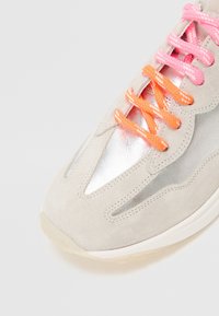 Light grey suede sneaker with shiny silver accents. Features contrasting pink and orange laces. Rounded toe and textured midsole.