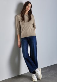Pull en tricot beige à manches longues associé à un jean large bleu foncé et des baskets blanches. Le modèle s'appuie contre un mur gris.