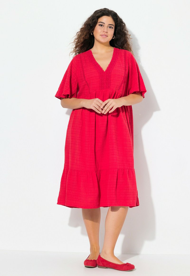 Robe en coton rouge avec des manches courtes à volants, décolleté en V et ourlet en niveaux. Portée avec des ballerines rouges assorties. Le tissu texturé ajoute de la profondeur.