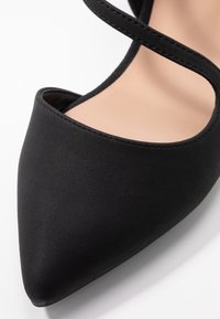 Zwarte stoffen hoge hak schoenen met een spitse neus en twee slanke kruisbanden. Gladde textuur met minimale stikseldetails.