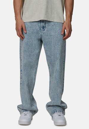 Mężczyzna ubrany w jasnoniebieskie, wzorzyste, szerokie spodnie jeansowe i białe trampki, częściowo widoczna jasnoszara, casualowa koszulka.