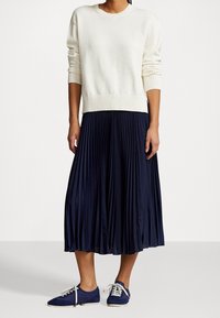 Suéter de punto crema con dobladillo acanalado, combinado con una falda midi plisada en azul marino y zapatillas azules con cordones y suelas blancas.