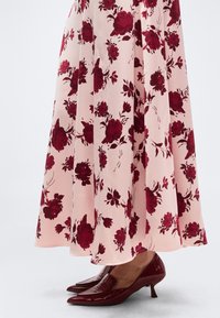 Ροζ φούστα με σκούρο κόκκινο λουλουδάτο σχέδιο, φορεμένη με λαμπερά σκούρα κόκκινα μυτερά παπούτσια με χαμηλό τακούνι, σε απλό ανοιχτό φόντο.