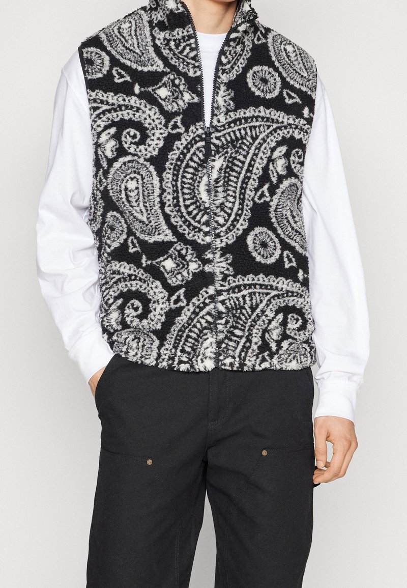 Homme portant un gilet polaire noir et blanc à motif cachemire sur une chemise blanche à manches longues et un pantalon noir, debout les mains dans les poches.