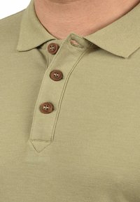 Khaki polo-shirts lavet af tekstureret stof med klassisk krave, tre brune knapper og en syet kantdetalje.