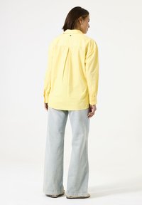 Camisa amarilla de manga larga con un pliegue en la parte posterior y detalle de botones, combinada con jeans de pierna ancha de color azul claro. La tela parece suave y texturizada.