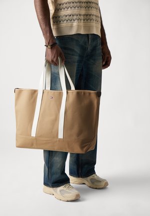 Sac fourre-tout beige avec des poignées blanches, fabriqué à partir d'un tissu résistant. Présente un logo discret à l'avant et une forme rectangulaire structurée.