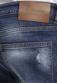 Jean en denim bleu avec une poche arrière comportant une petite pièce usée et une étiquette en cuir marron cousue indiquant "ZS PREMIUM."