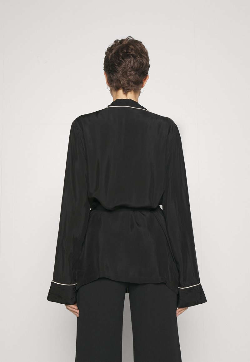 Zwarte satijnen blouse met lange mouwen, een ceintuur in de taille en witte piping op de kraag en manchetten. Gladde textuur met een comfortabele pasvorm.