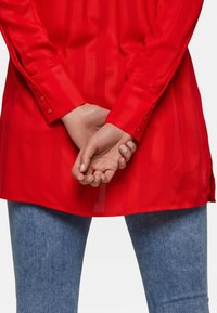 Camicia rossa a maniche lunghe con righe verticali, polsini abbottonati e orlo curvo, indossata sopra un denim azzurro chiaro. Mani incrociate dietro la schiena.
