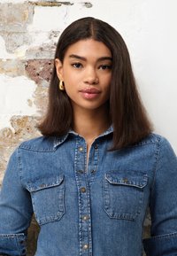 Junge Frau mit glatten dunklen Haaren trägt goldene Creolen und ein blaues Jeanshemd vor einer abgenutzten weißen Ziegelwand.