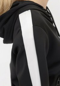 Haut à capuche noir avec une bande blanche le long de l'épaule. Fabriqué à partir d'un matériau lisse et extensible, avec une fermeture éclair à l'avant et une capuche ajustable.