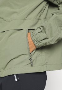 Main dans la poche zippée d'une veste vert clair avec un poignet élastique, sur un pantalon noir avec un texte blanc partiel visible.
