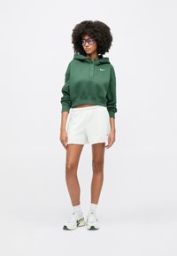 Sweat à capuche court vert avec fermeture à boutons et logo, associé à un short gris clair et des baskets blanches avec des accents verts.