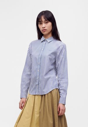 Jeune femme aux longs cheveux foncés portant une chemise boutonnée à rayures bleues et blanches et une jupe plissée beige sur un fond uni.