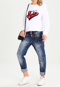 Vit sweatshirt med röd glittertext "Hola," kombinerad med slitna blå jeans som är uppvikta vid bensluten och vita sneakers.