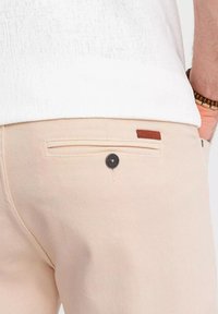 Pantalons en coton beige avec une poche arrière dotée d'un bouton noir et d'un accent en cuir brun. Le tissu a une texture lisse.