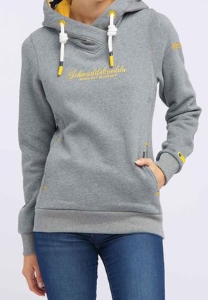 Vrouw die een grijze hoodie draagt met gele uiteinden van de trekkoorden en letters, een blauwe spijkerbroek, en haar hand in de voorkantzak.