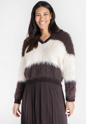 Vrouw met lang donker haar lacht, ze draagt een zachte V-hals trui met witte en donkerbruine horizontale strepen en een geplooide donkere rok.