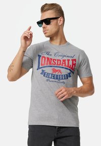 Grå bomulds t-shirt med et rød- og blåt grafisk print med "Lonsdale" tekst og et løvedesign. Rund hals, korte ærmer, ensartet tekstur.