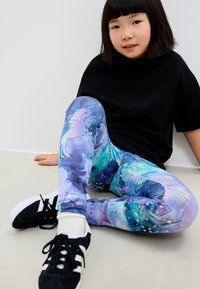 Färgglada leggings med enhörningsmotiv i lila, blått och teal med en glansig yta, kombinerade med en svart tröja och svarta sneakers.