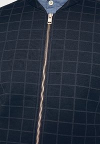 Giacca blu scuro con motivo a griglia grigio chiaro, cerniera argentata al centro, indossata sopra una camicia blu con un bottone bianco.