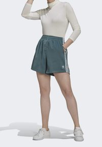 Tealfärgade atletiska shorts med lös passform, elastisk midja och sidofickor. Har tre vita ränder längs sidorna och en Adidas-logotyp.